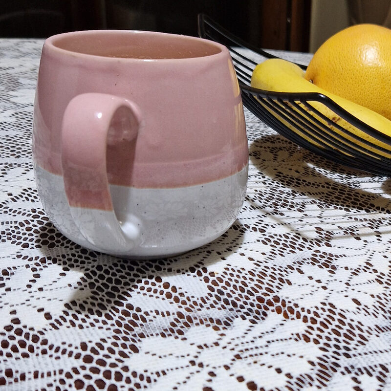 Foto real hecha con el móvil: taza rosa y blanca junto a un frutero, sobre un mantel de encaje